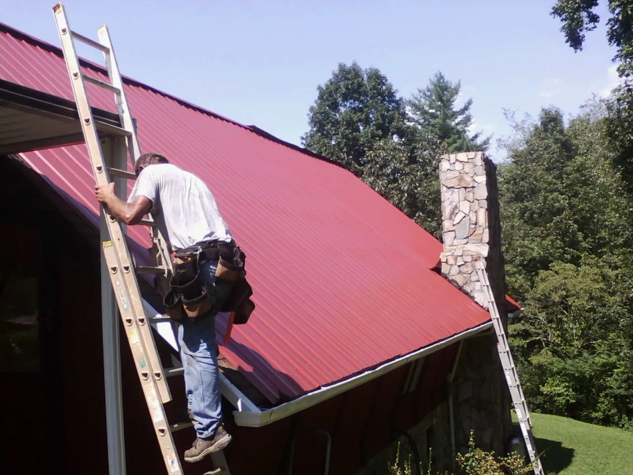 Roofer on a ladder beside a red metal roof and stone chimney — Soffit & Fascia Repair by Roofing Brothers in Opp